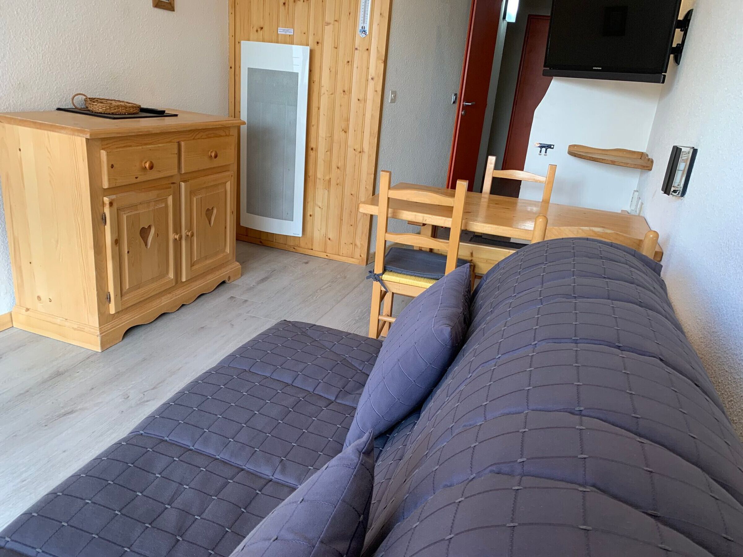 Une pièce comprenant un lit, une commode en bois, une chaise, une petite table et un téléviseur fixé au mur.