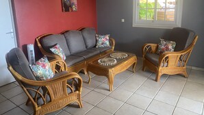 A room with a red wall, a grey sofa, two wicker chairs, a small table with decorative items, and a window with a view of greenery. - Villa for 6 people, swimming pool, sea view, close to Sainte Anne beaches (SAINTE ANNE)