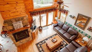 Interior acolhedor de uma cabana com lareira de pedra, sofá de couro e uma grande janela com vista para um deck.
