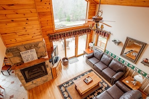 A cozy cabin interior with a stone fireplace, leather sofa, and a large window overlooking a deck.