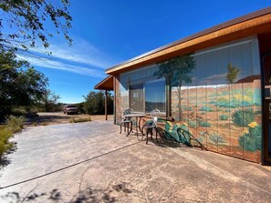 A modern building with a patio area featuring a table and chairs, a large window with a view of the desert landscape, and a mural on the wall.