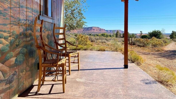 A covered outdoor area with two wooden chairs and a concrete floor, overlooking a desert landscape with sparse vegetation and distant mountains.