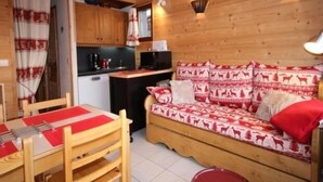 A cozy cabin interior with a wooden dining table, a sofa with patterned cushions, and a kitchen area. - Cozy apartment for 5, south balcony, near slopes (Les Saisies)