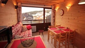 A cozy room with a wooden interior, a dining table with red placemats, a sofa with patterned cushions, and a view of a town through large windows. - Cozy apartment for 5, south balcony, near slopes (Les Saisies)