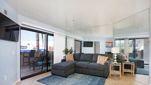 Una sala moderna con un sofá modular, un ventanal que da a un balcón y un televisor de pantalla plana montado en la pared.