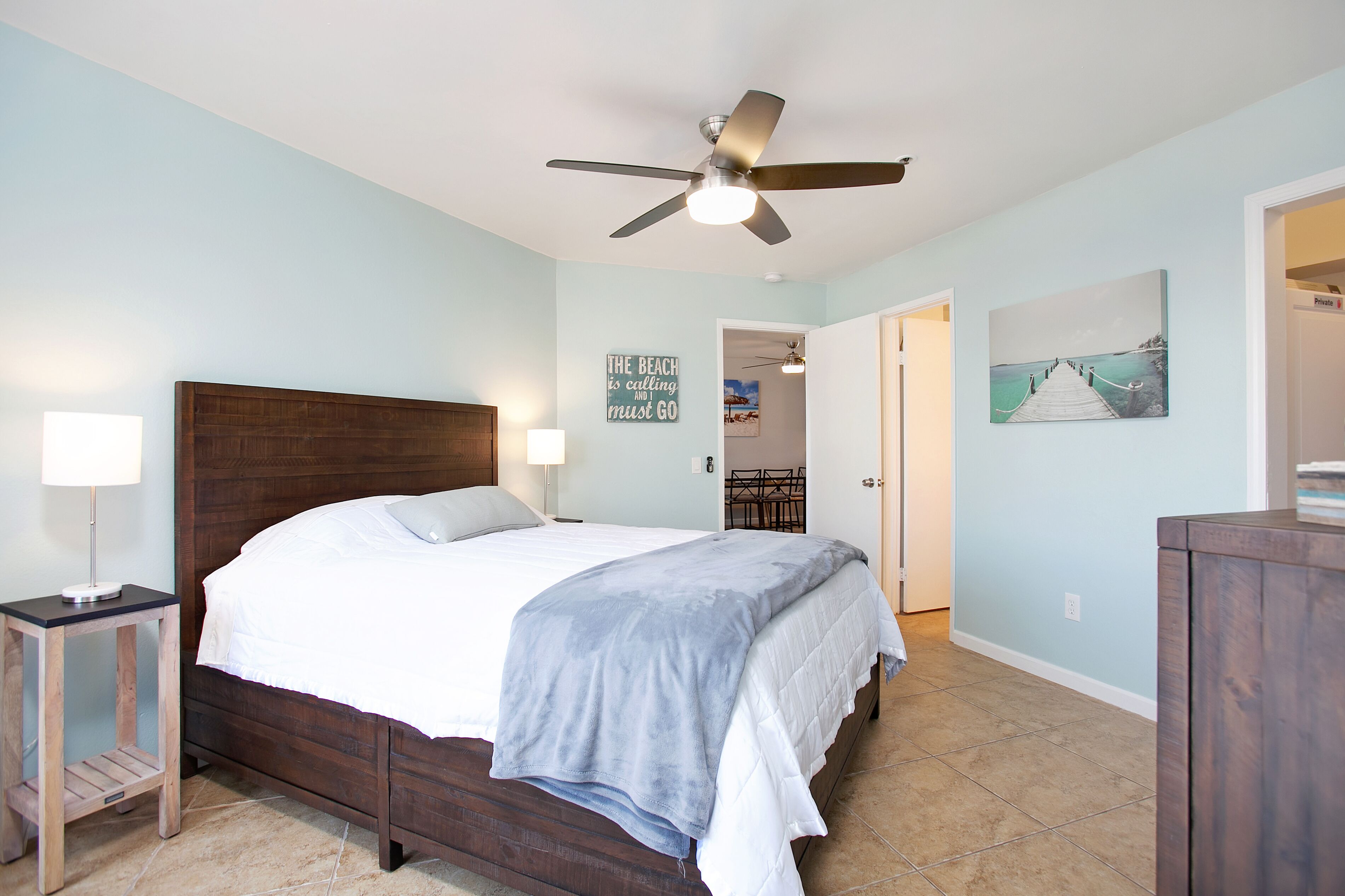 A bedroom with a bed, a ceiling fan, a nightstand, a lamp, a painting, and a door leading to another room.
