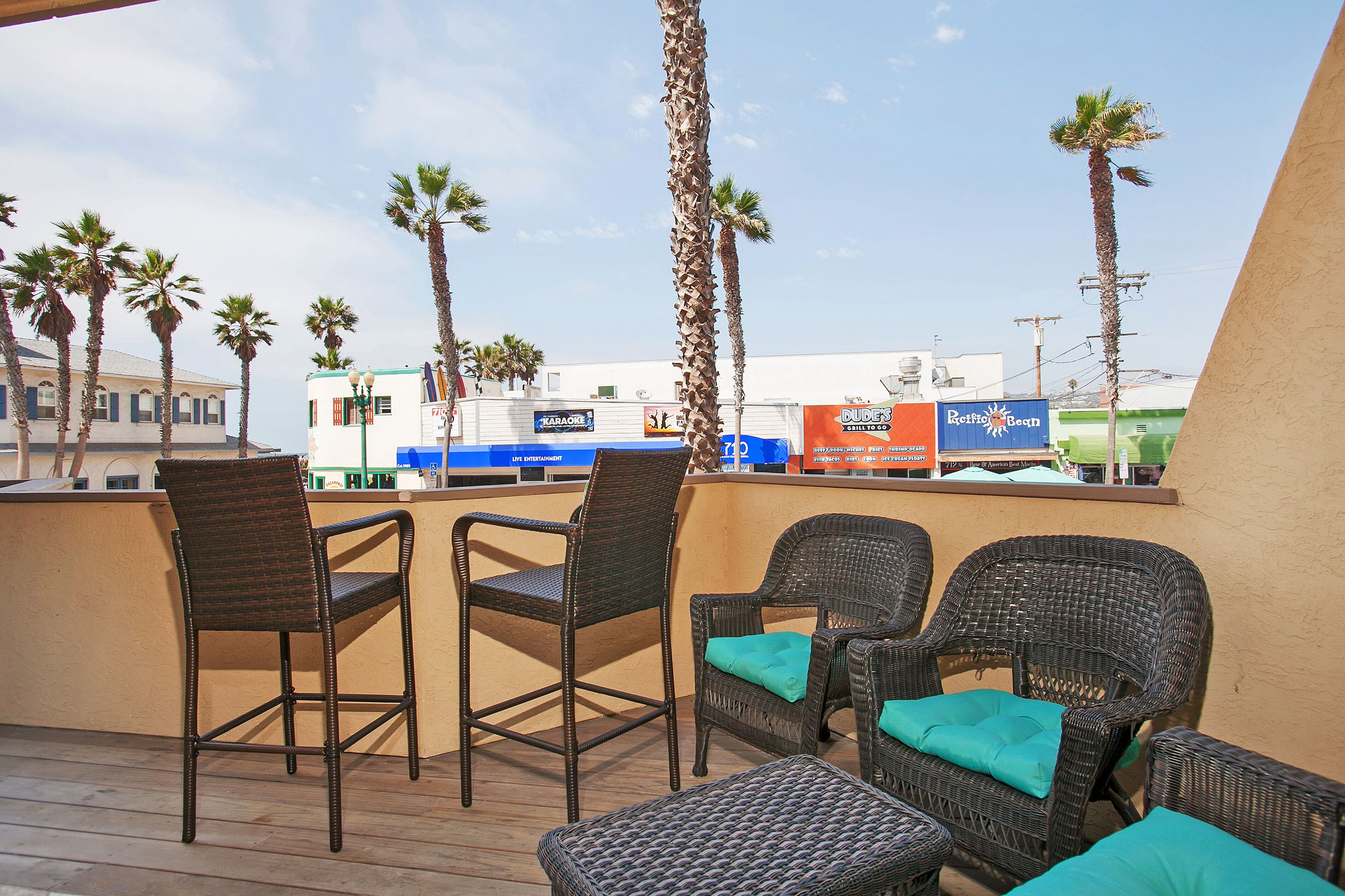 A deck with wicker chairs and a bar area, overlooking a beach with palm trees and a building with signs.