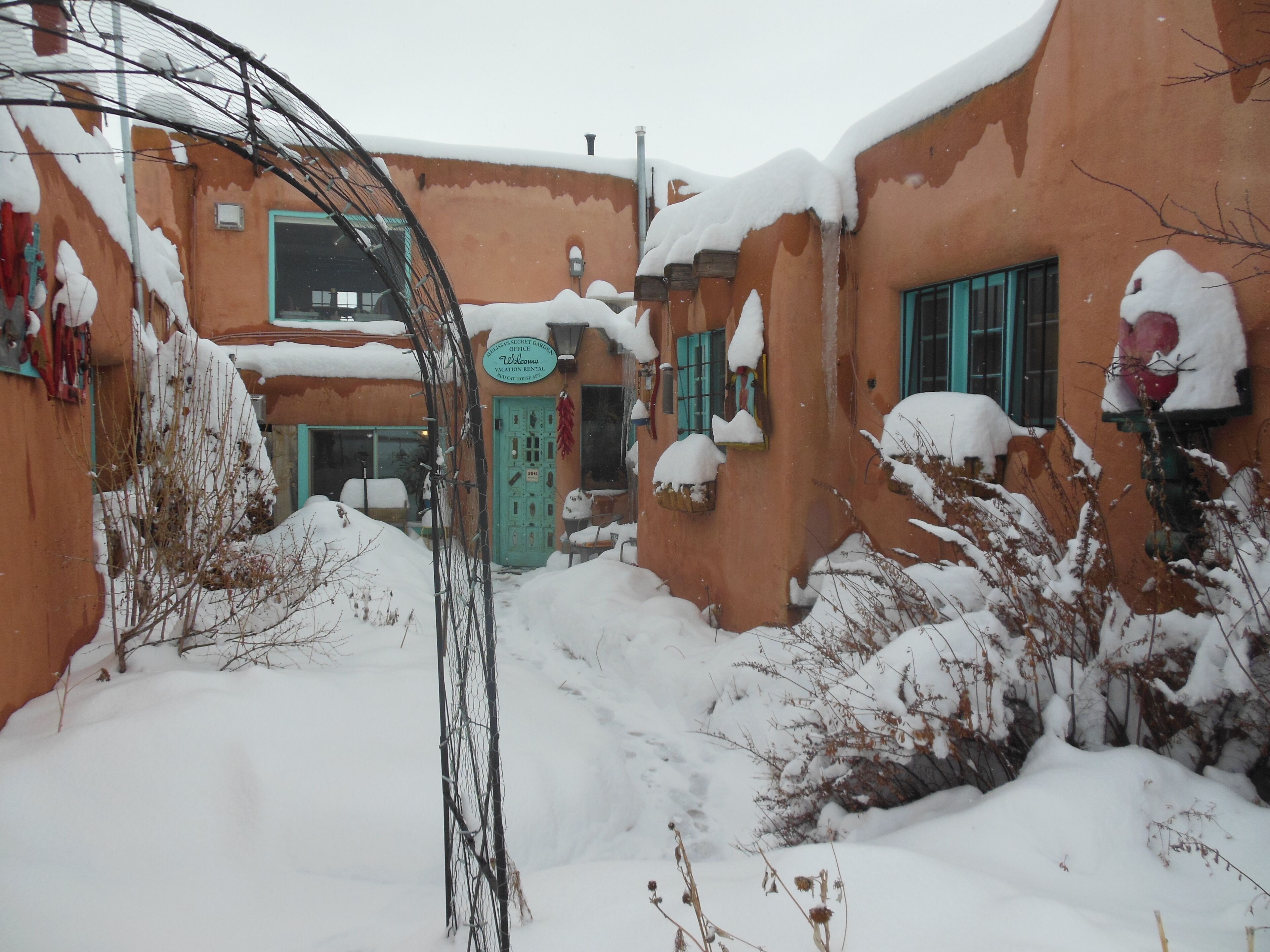 A snow-covered courtyard with a building featuring a turquoise door and a sign that reads 'Welcome.'
