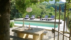 Un área junto a la piscina con sillones de descanso y una mesa, rodeada de árboles y vistas a un paisaje montañoso.