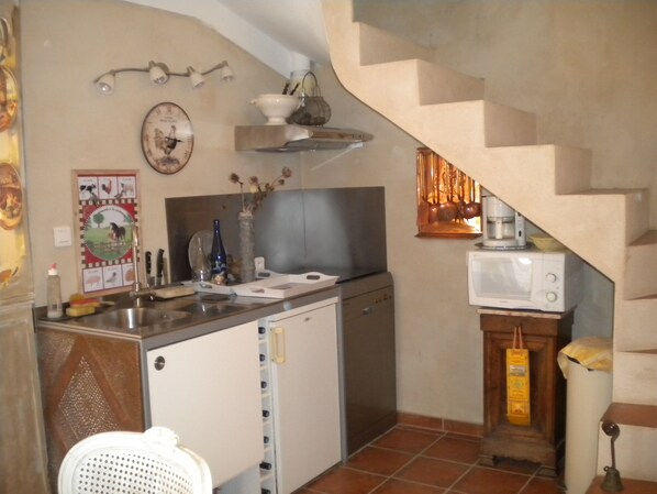 Cocina compacta con lavamanos, estufa y microondas. Hay un refrigerador pequeño y un armario de madera. Un reloj está montado en la pared, y un florero con flores secas está sobre la encimera.