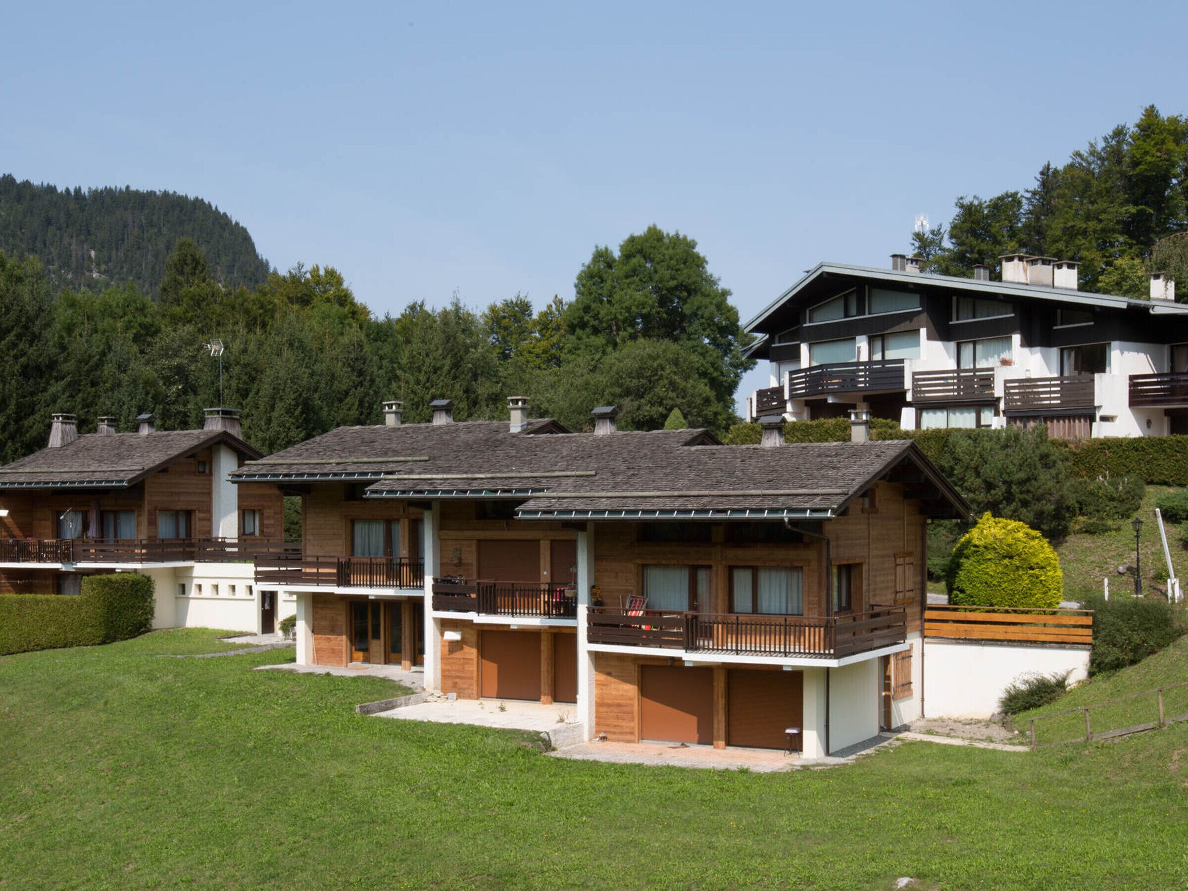 A row of chalet-style buildings with balconies, set against a backdrop of trees and a mountain.