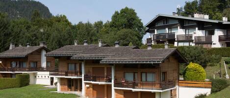 A row of chalet-style buildings with balconies, set against a backdrop of trees and a mountain.