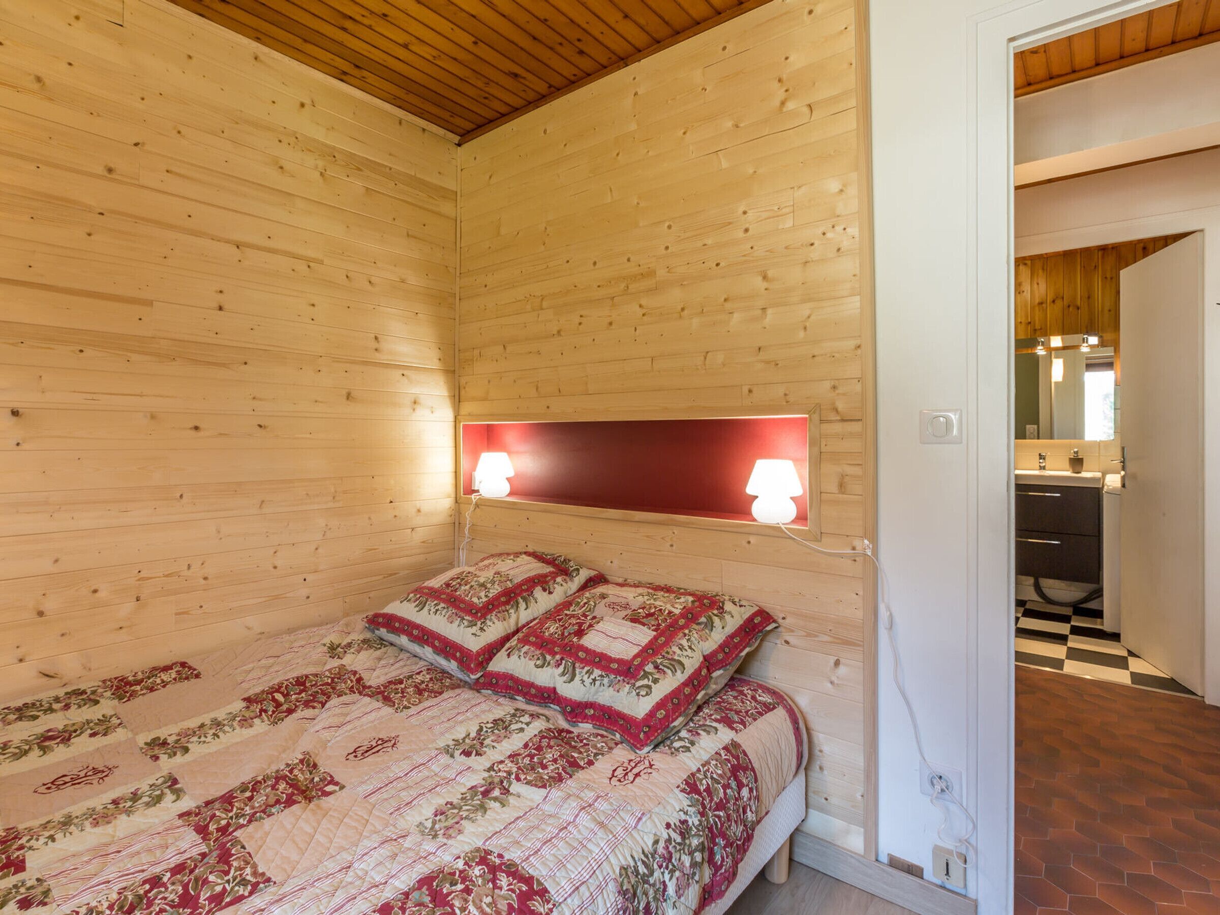 A wooden cabin room with a bed, two bedside lamps, and a visible bathroom with a checkered floor.