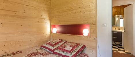 A wooden cabin room with a bed, two bedside lamps, and a visible bathroom with a checkered floor.