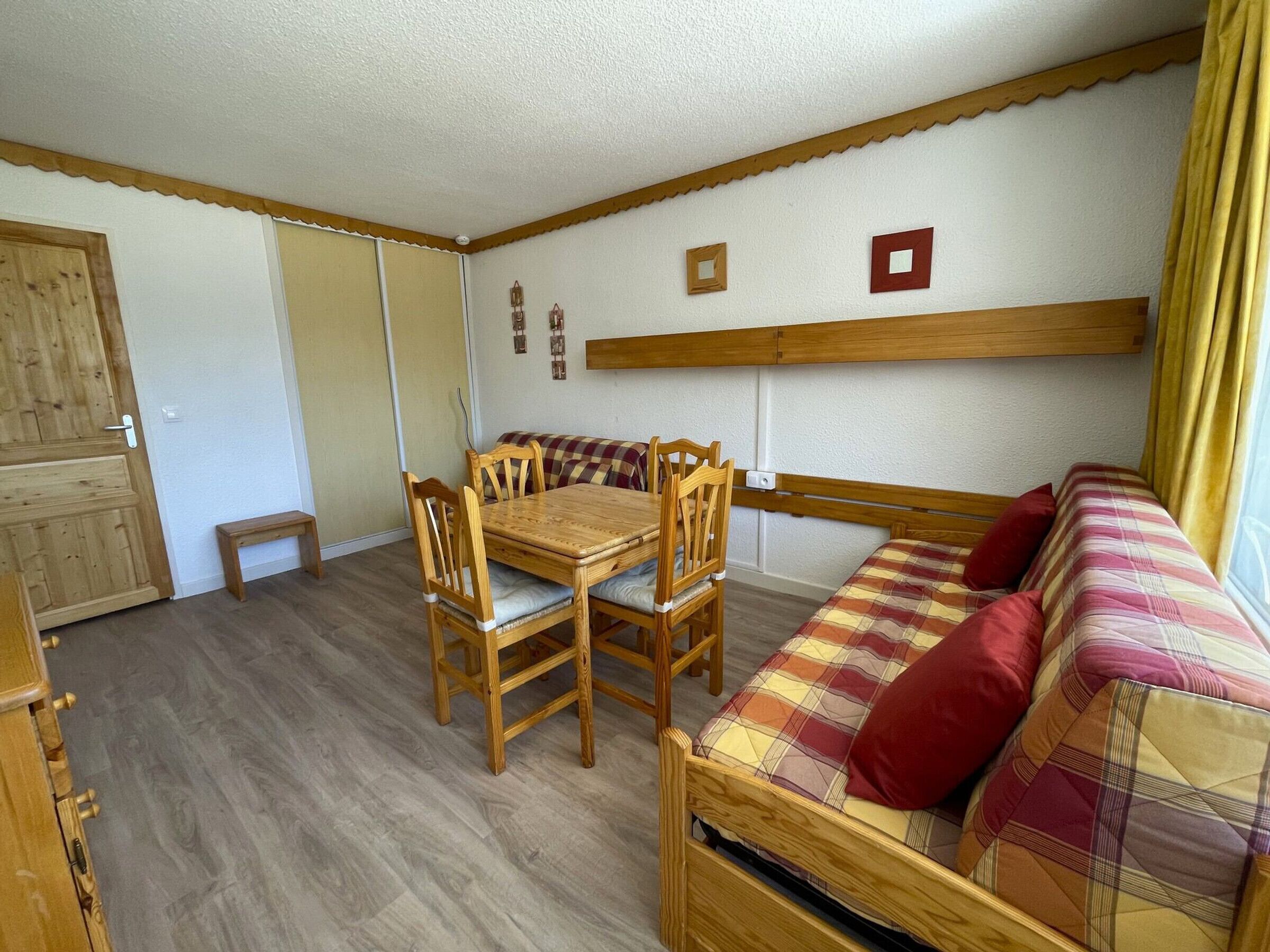 A compact room with a wooden dining table and chairs, a sofa with a plaid blanket, and a wooden cabinet.
