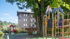 Leikkikenttä, jossa on kiipeilytelineitä ja liukumäki, ja se on ympäröity puilla. Kentän läheisyydessä on myös parvekkeellinen rakennus.
