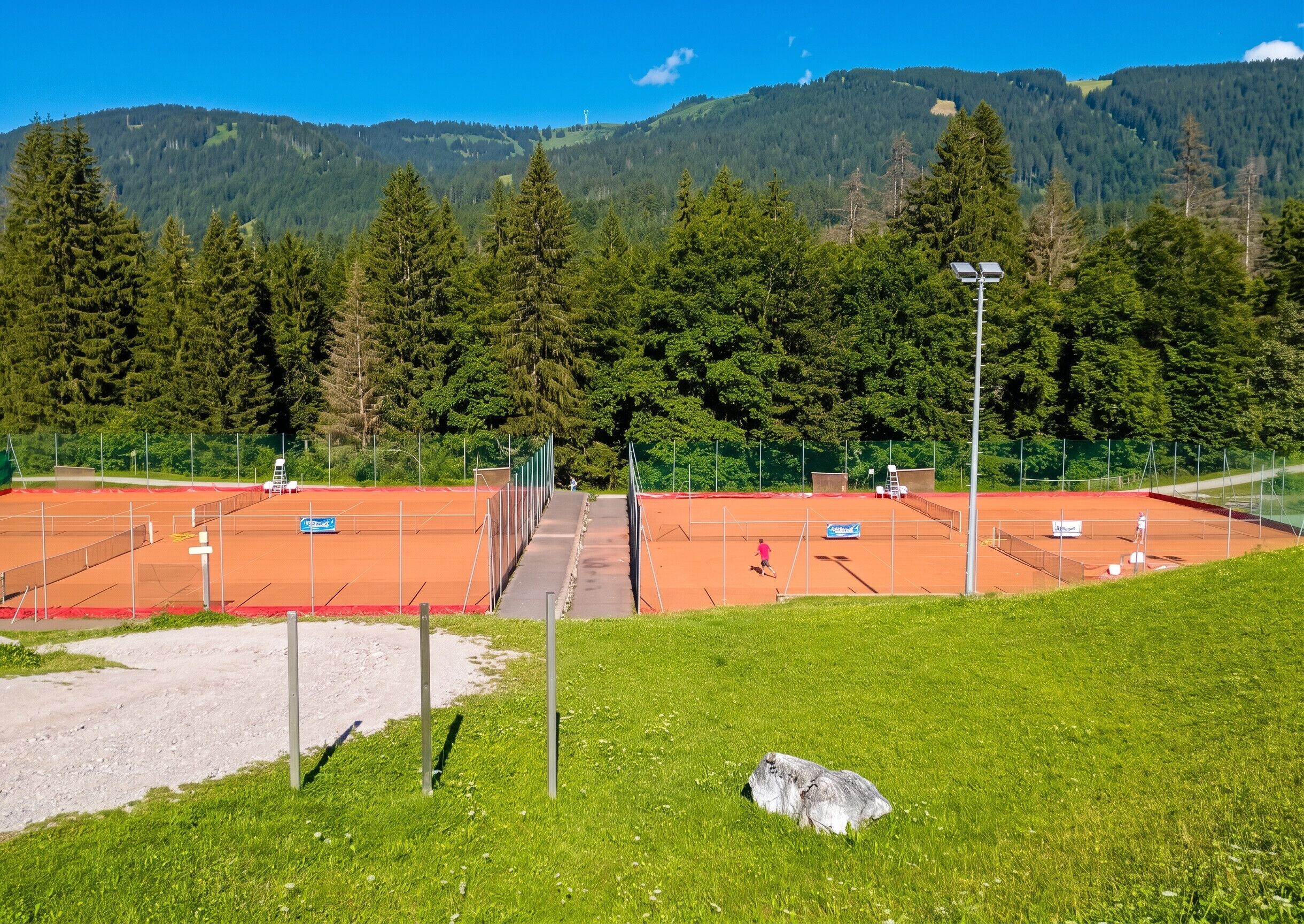 Cancha de tenis con red, rodeada por una valla y con montañas al fondo.