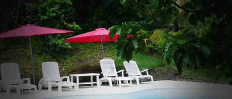 Un espace piscine agrémenté de chaises longues blanches et de parasols rouges, le tout entouré d’une végétation luxuriante.