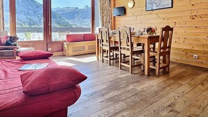 A cozy room with a wooden interior, a dining table with chairs, a sofa with red cushions, and a large window offering a mountain view.
