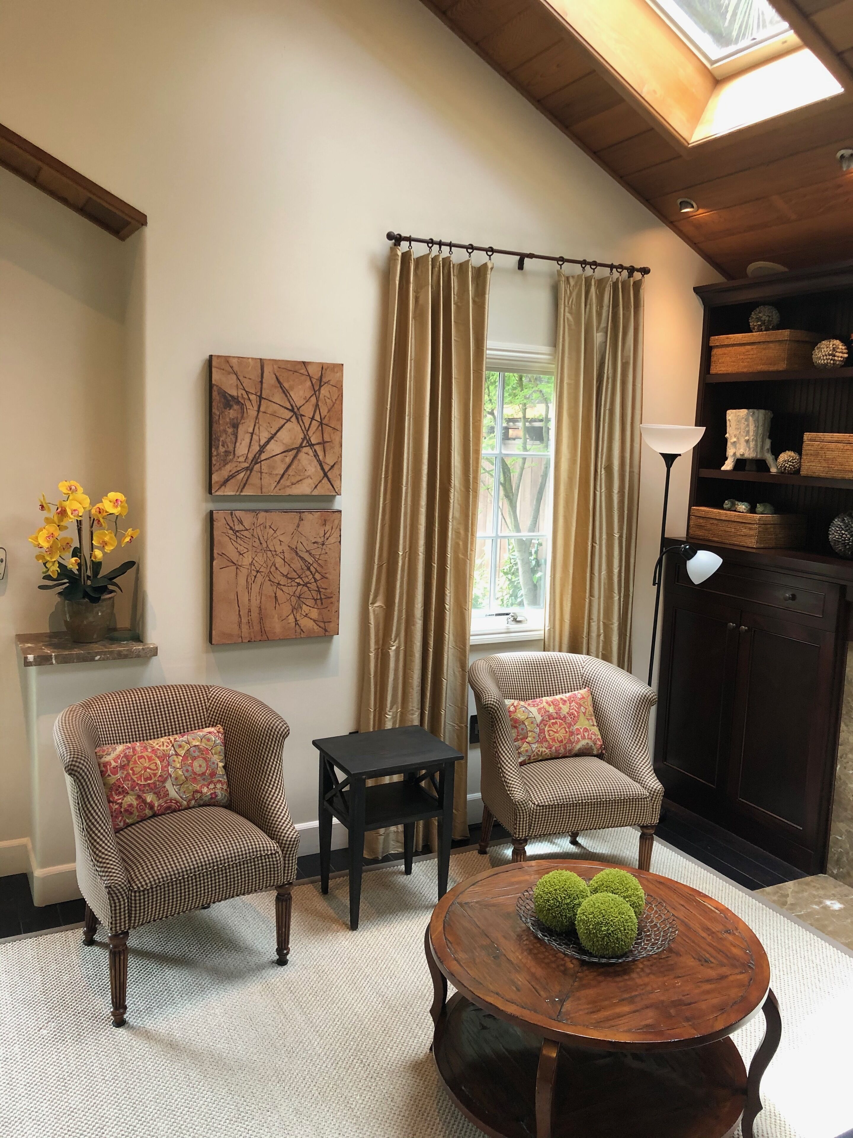 A cozy living room with two armchairs, a small table, and a round coffee table with decorative balls. There are two abstract paintings on the wall, a window with curtains, and a bookshelf with decorative items.