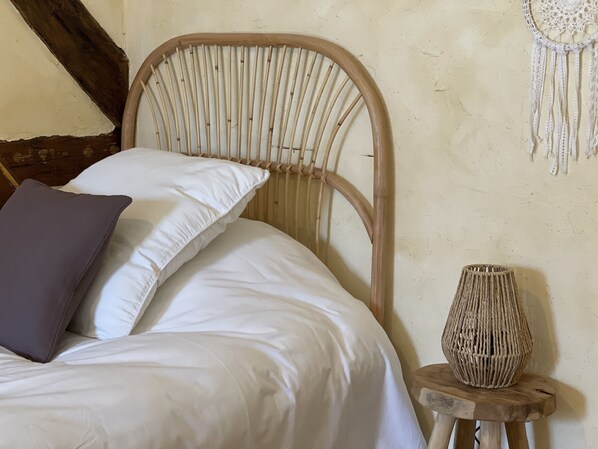 A bed with white bedding and a single dark pillow, a woven vase on a small wooden stool, and a wall with a simple, textured design.