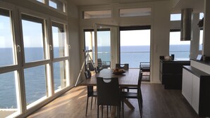 Une cuisine moderne avec vue sur la mer, un coin repas avec une table et des chaises, et une grande fenêtre équipée d’un trépied.