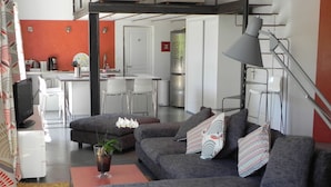 A modern living room with a grey sofa, a coffee table, and a plant. There is a kitchen area in the background with white cabinets and a refrigerator. - A PEACEFUL HEAVEN IN THE HEART OF LUBERON (Cucuron)