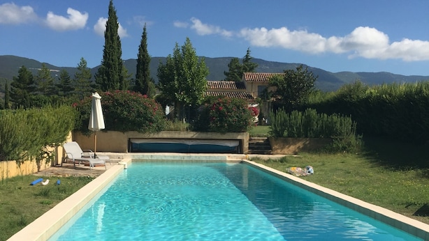 A rectangular swimming pool with clear blue water, surrounded by a grassy area, a few trees, and a building with a tiled roof in the background. - A PEACEFUL HEAVEN IN THE HEART OF LUBERON (Cucuron)