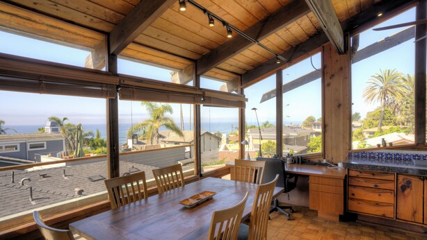 Un comedor espacioso con una mesa y sillas de madera, ventanas amplias con vistas al océano y palmeras, y una cocina con armarios de madera y una encimera.
