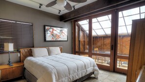 Un dormitorio con una cama grande, una mesita de noche, una lámpara, una obra de arte enmarcada y una puerta corrediza de vidrio que da a un balcón.
