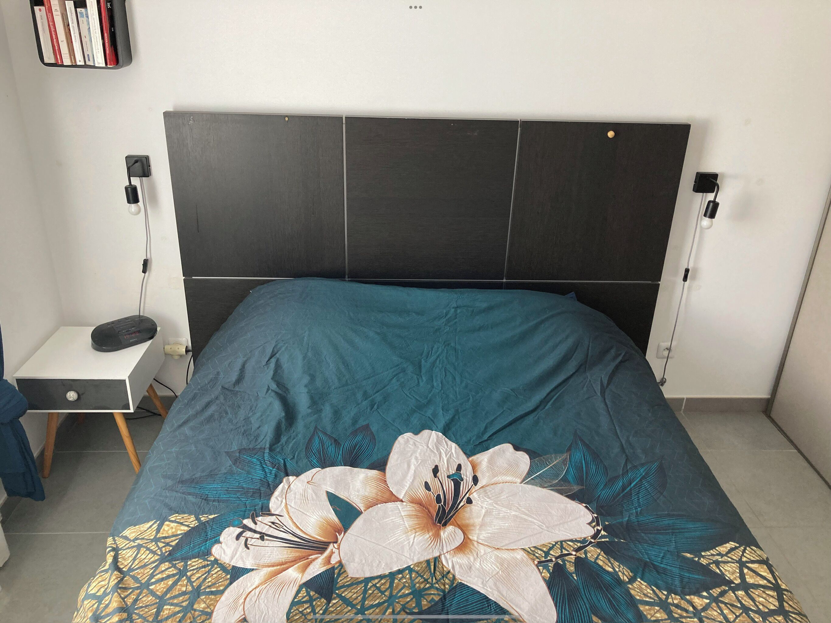 A bedroom with a bed, a bedside table, a nightstand, a wall-mounted shelf with books, and a power outlet.