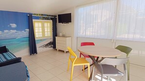 A room with a beach mural, a TV, a dining table with chairs, and a window with curtains. - GITE FOR 4 PERS WITH POOL (Agde)