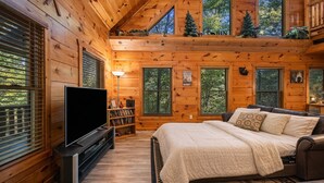 Une chambre dans un chalet en rondins, avec un lit, une télévision, une bibliothèque et une fenêtre sur laquelle est affiché un panneau indiquant « CHALET ».