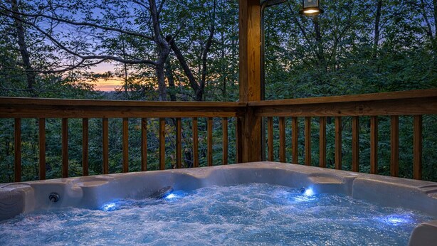 Un bain à remous sur une terrasse en bois, avec vue sur les arbres et un coucher de soleil en arrière-plan.