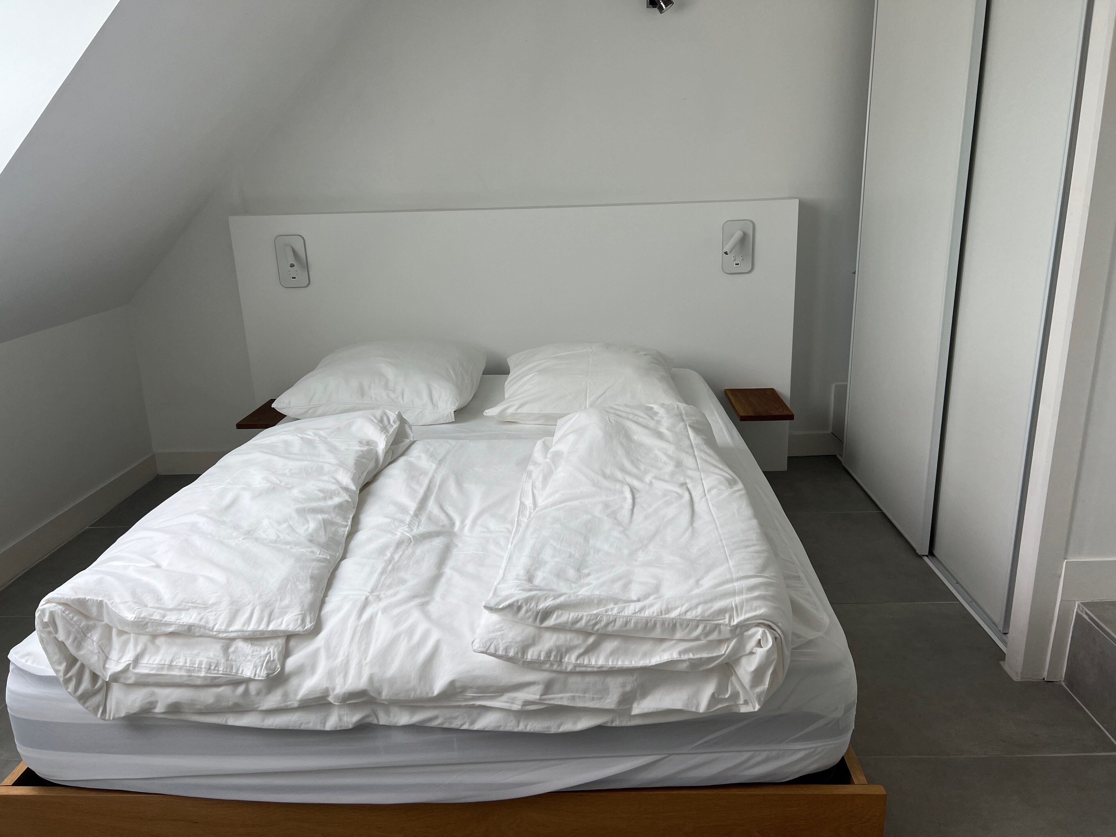 A neatly made bed with white bedding in a room with white walls and a sloped ceiling.