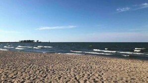Une plage de sable avec des vagues, un ciel dégagé et une côte lointaine parsemée de bâtiments.