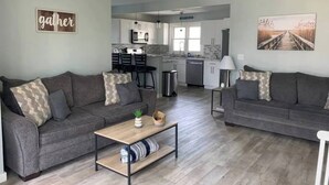 Un salon moderne avec deux canapés gris, une table basse en bois et une cuisine en arrière-plan.