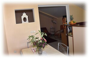 A dining area with a table, chairs, a vase with flowers, a wall-mounted shelf with a decorative item, and a kitchen area with a fruit bowl.