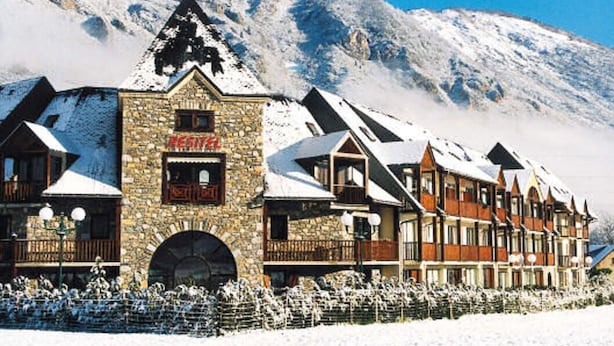 Un hotel de montaña cubierto de nieve con fachada de piedra, varios balcones y un telesilla al fondo.