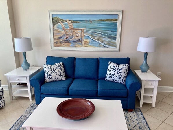 A living room with a blue sofa, two matching lamps, a painting of a beach scene, and a white coffee table with a bowl. - 🏖️Aqua 1410 Oceanfront; King/Dbl beds,  2 full baths (Panama City Beach)