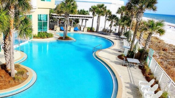 A curved pool with a clear blue water, surrounded by palm trees and lounge chairs, with a beach and ocean view in the background.
