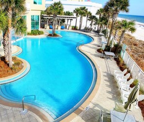 A curved pool with a clear blue water, surrounded by palm trees and lounge chairs, with a beach and ocean view in the background. - 🏖️Aqua 1410 Oceanfront; King/Dbl beds,  2 full baths (Panama City Beach)
