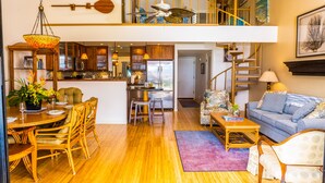 A spacious open-plan living area with a dining table, chairs, and a sofa. A kitchen is visible in the background with modern appliances and wooden cabinets. - Ma'alaea Surf, Unit G9-2BR Beachfront Condo, AC, Wi-Fi, TV, Pool, Kitchen (Kihei)