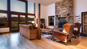 Un vaste salon doté d’une cheminée en pierre, de grandes fenêtres offrant une vue sur les montagnes et d’un coin salon confortable avec un fauteuil et un canapé en cuir.