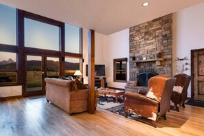 A spacious living room with a stone fireplace, large windows offering a view of the mountains, and a cozy seating area with a leather armchair and sofa. - 6-BR Teton View Ranch with Hot Tub (Driggs)