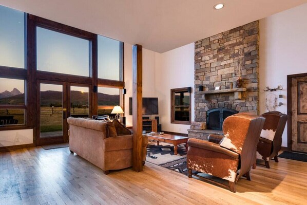 A spacious living room with a stone fireplace, large windows offering a view of the mountains, and a cozy seating area with a leather armchair and sofa.