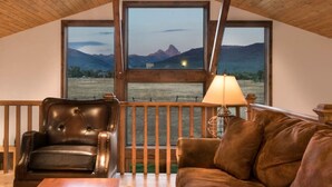 Une pièce chaleureuse avec un plafond en bois, une grande fenêtre donnant sur les montagnes, un fauteuil en cuir, un canapé marron et une table avec une lampe.