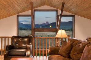 A cozy room with a wooden ceiling, a large window offering a mountain view, a leather armchair, a brown sofa, and a table with a lamp. - 6-BR Teton View Ranch with Hot Tub (Driggs)