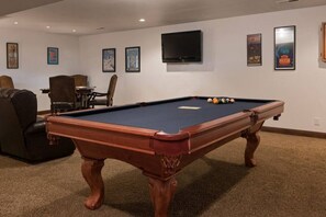 A pool table in a room with a flat-screen TV mounted on the wall, framed artwork on the walls, and a brown leather sofa.