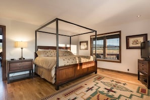 Uma cama de dossel com estrutura de madeira e colcha estampada, uma mesa de cabeceira com abajur, um aparador e uma janela grande com persianas.